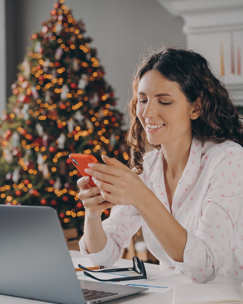 A woman sitting in front of a Christmas tree with her phone and her laptop